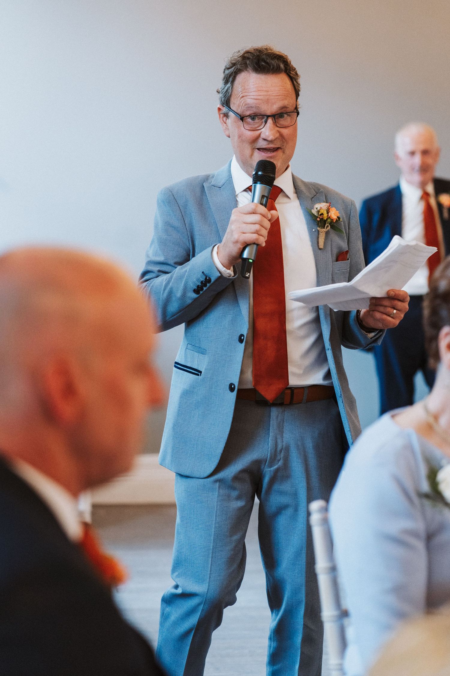 A speaker in a light blue suit delivers a speech at a formal event while holding papers.