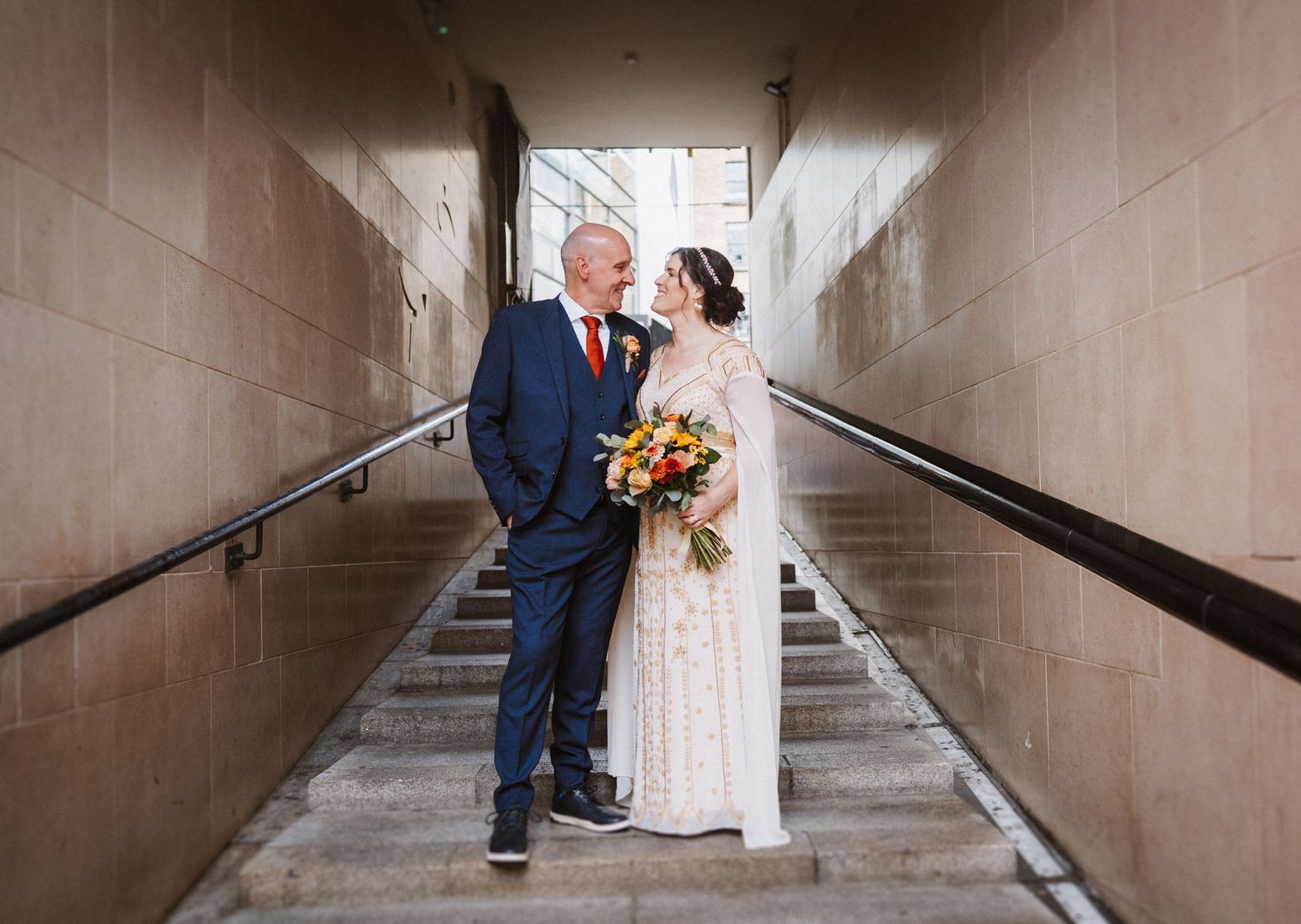 A couple shares an intimate moment on a staircase in a tan corridor while holding a vibrant yellow floral bouquet.