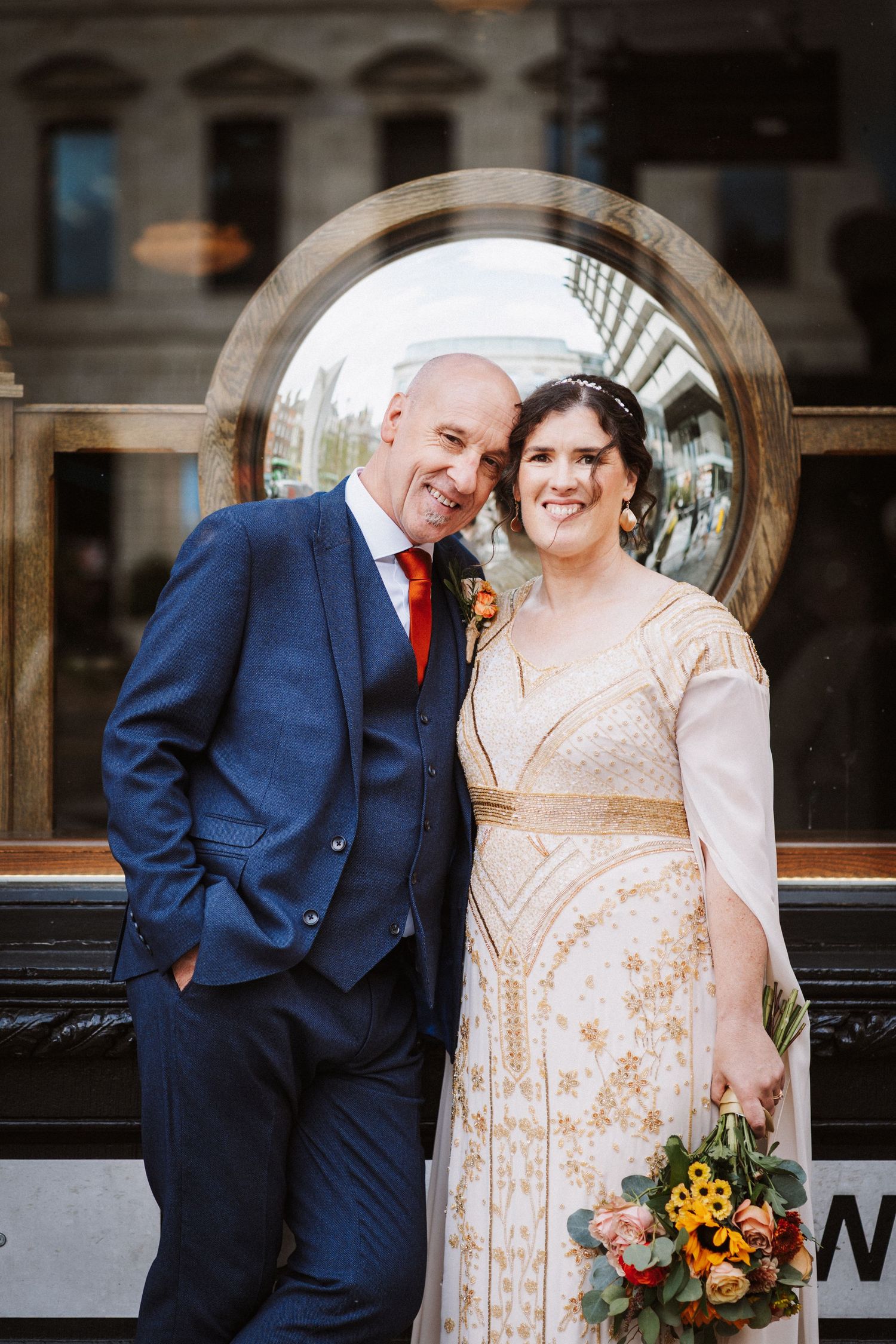 A couple in navy suit and gold embroidered dress pose together in a romantic series of loving embraces by a window.
