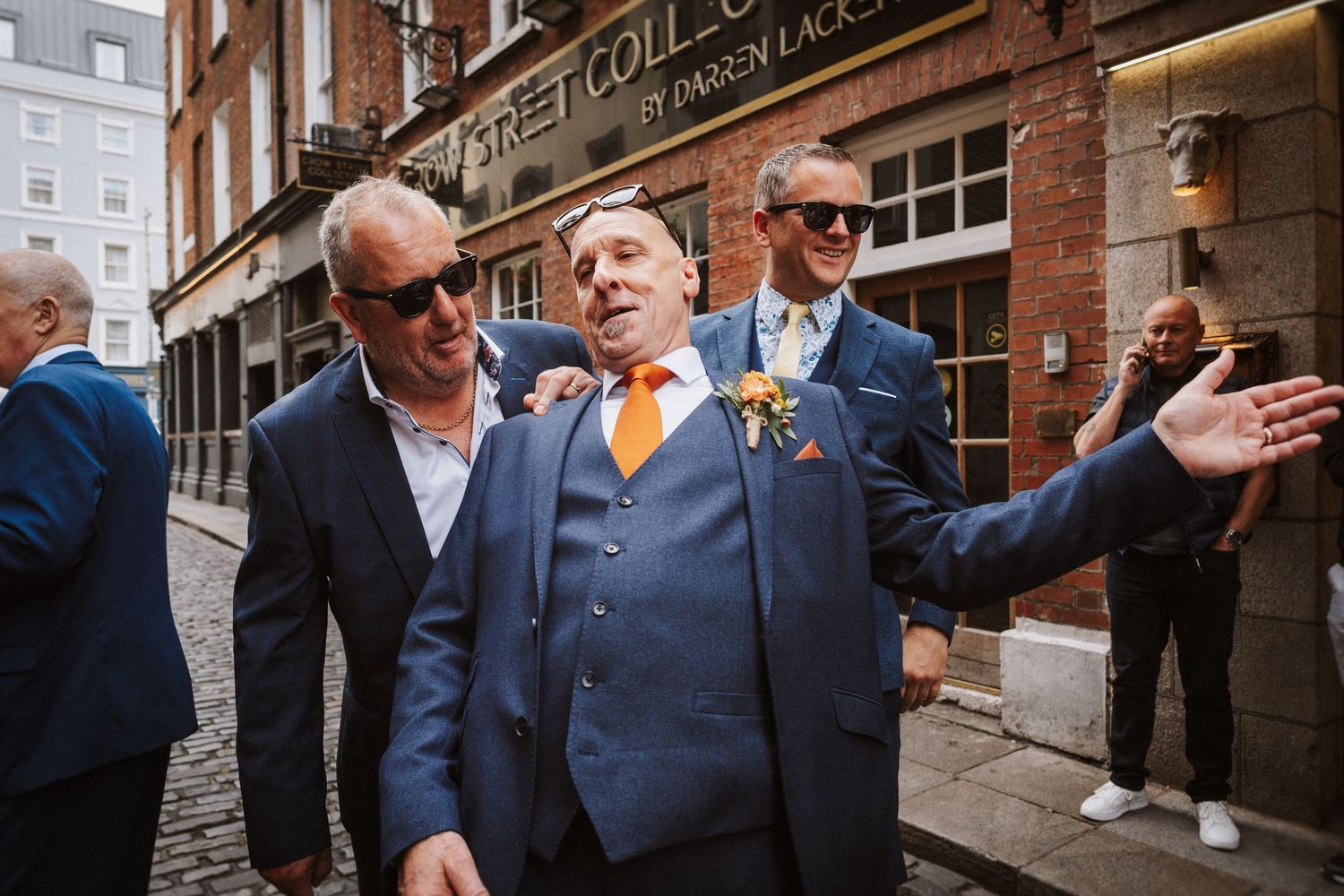 Wedding party guests interact outside a brick building with classic architecture on a sunny day.
