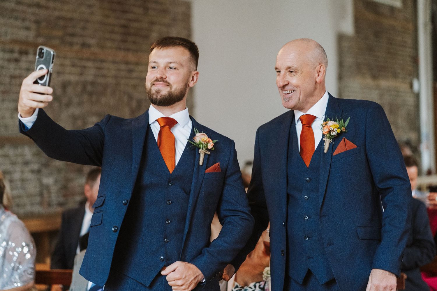 Groomsmen in navy blue suits with orange accents share a laugh during a wedding celebration in a brick-walled venue.