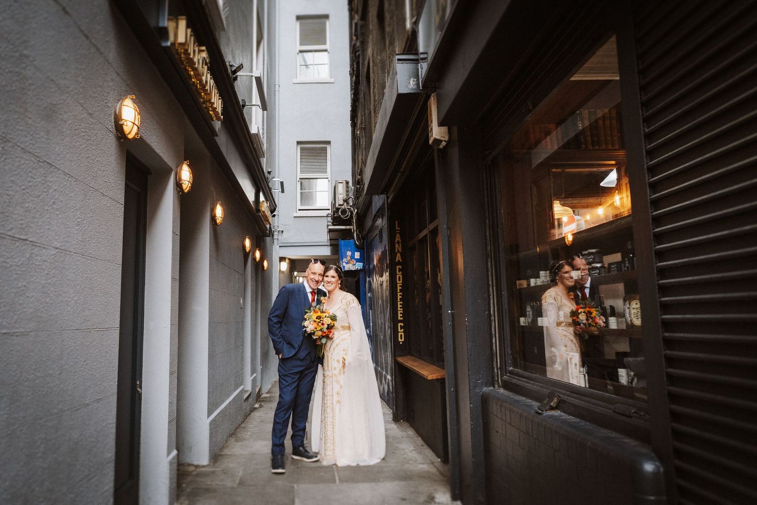 Two people in formal wedding attire embrace in a narrow urban alleyway with soft lighting and shop windows.