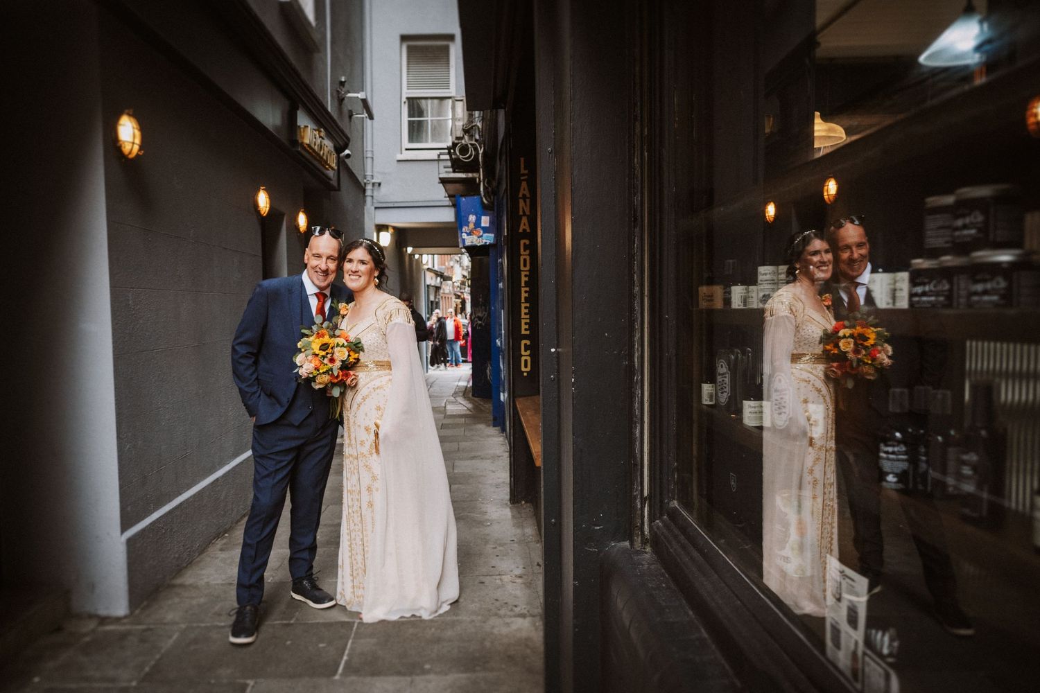 A couple shares romantic moment in urban alleyway with their reflection visible in storefront window.