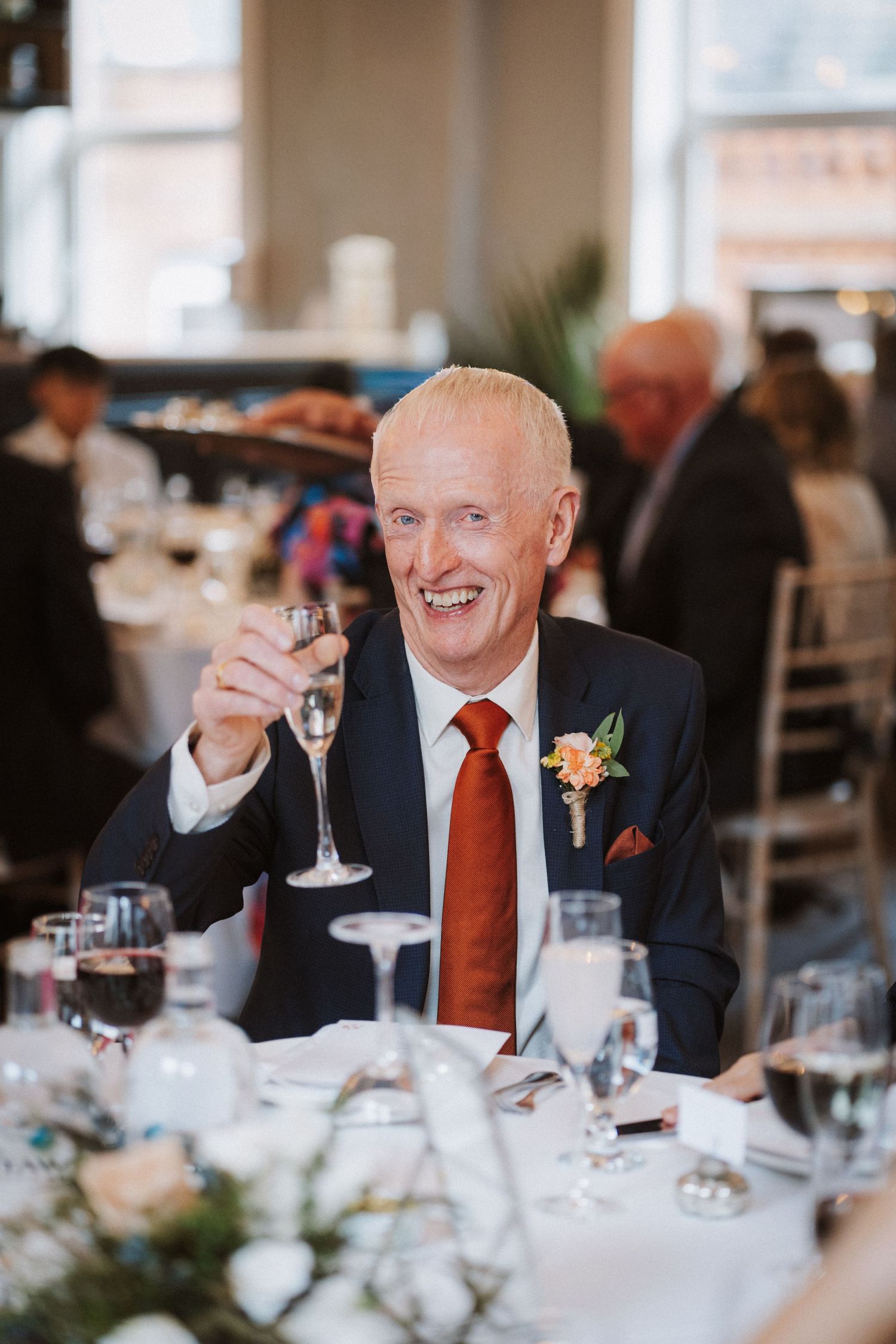 Wedding reception guests in navy suits and red ties smile while raising glasses in a toast at an elegant table setting.