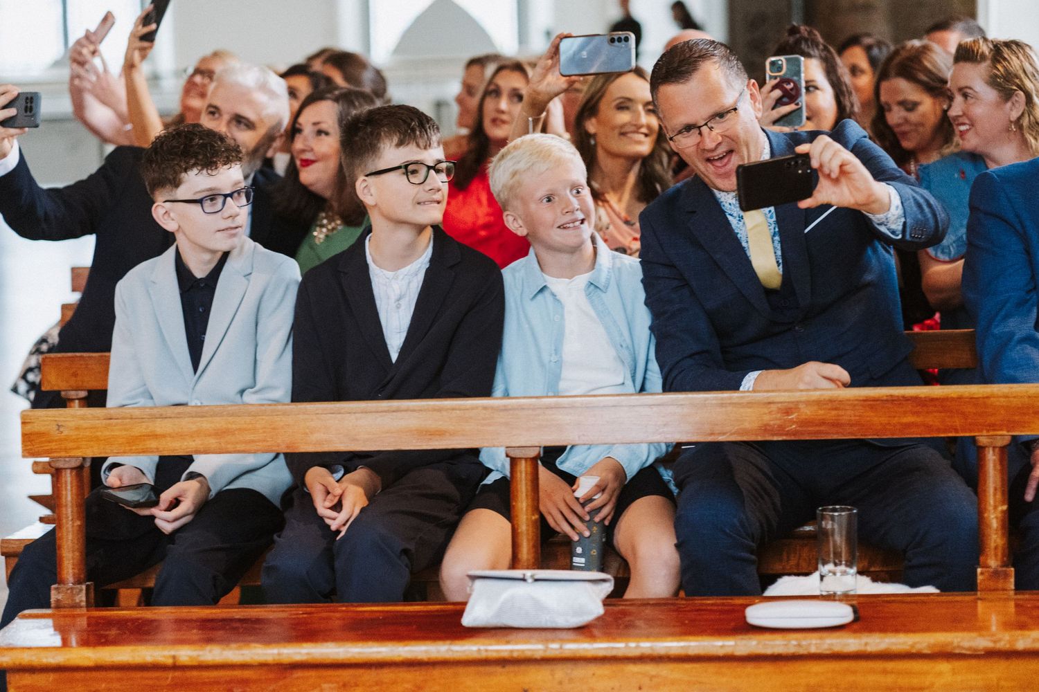 A crowd of excited spectators on wooden benches take selfies and photos during an outdoor event.