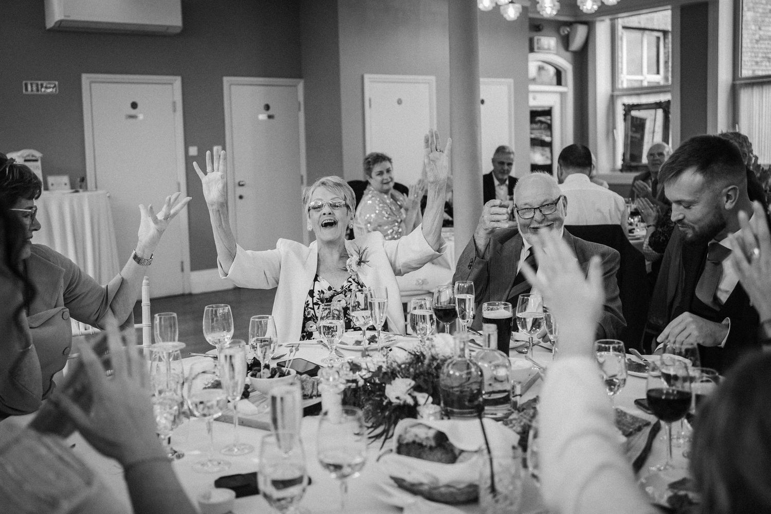 Black and white image of a wedding reception dining venue with tables, place settings and guests enjoying the celebration.