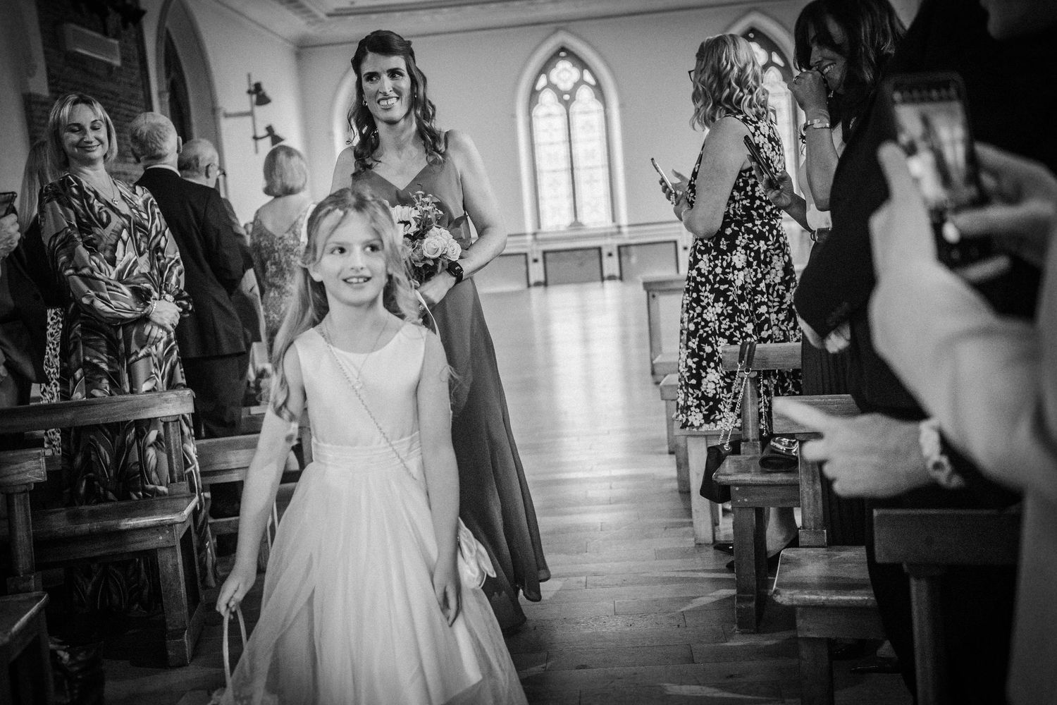 Black and white wedding scene in a church hall with pews and arched windows shows a young attendant in white dress.