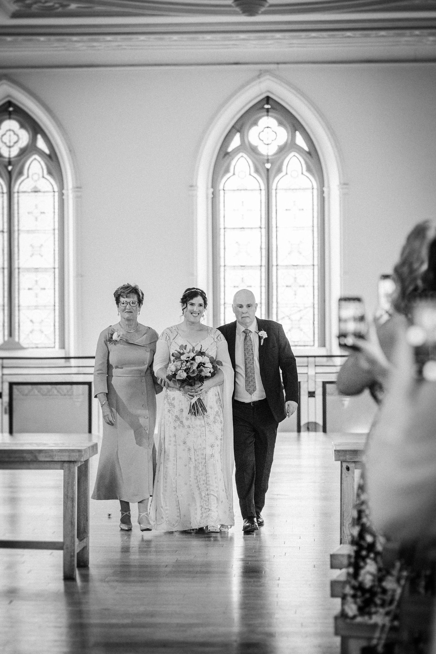 Black and white sequence of figures walking down a church aisle with gothic arched windows in the background.