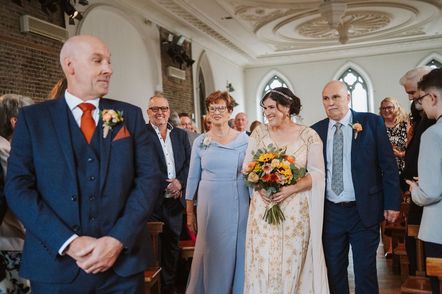 Wedding guests gather in an ornate church interior with elegant white molding and decorative details on the ceiling.
