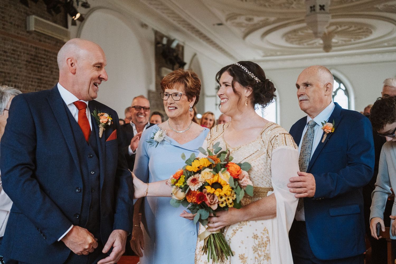A wedding celebration showing people dancing and embracing with ornate white molded ceiling details above.