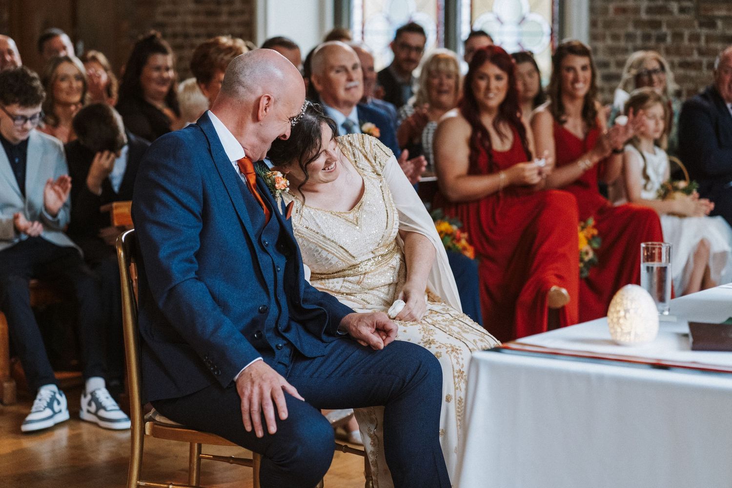 Smiling couple shares an intimate moment during their wedding ceremony while seated in front of brick wall.