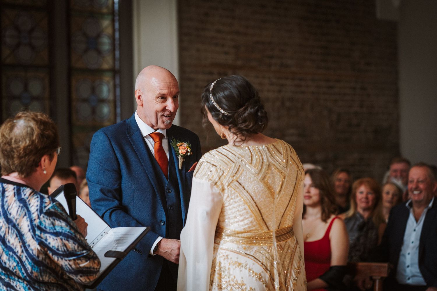 Wedding ceremony taking place in a brick-walled venue with guests seated in the background and participants standing at front.