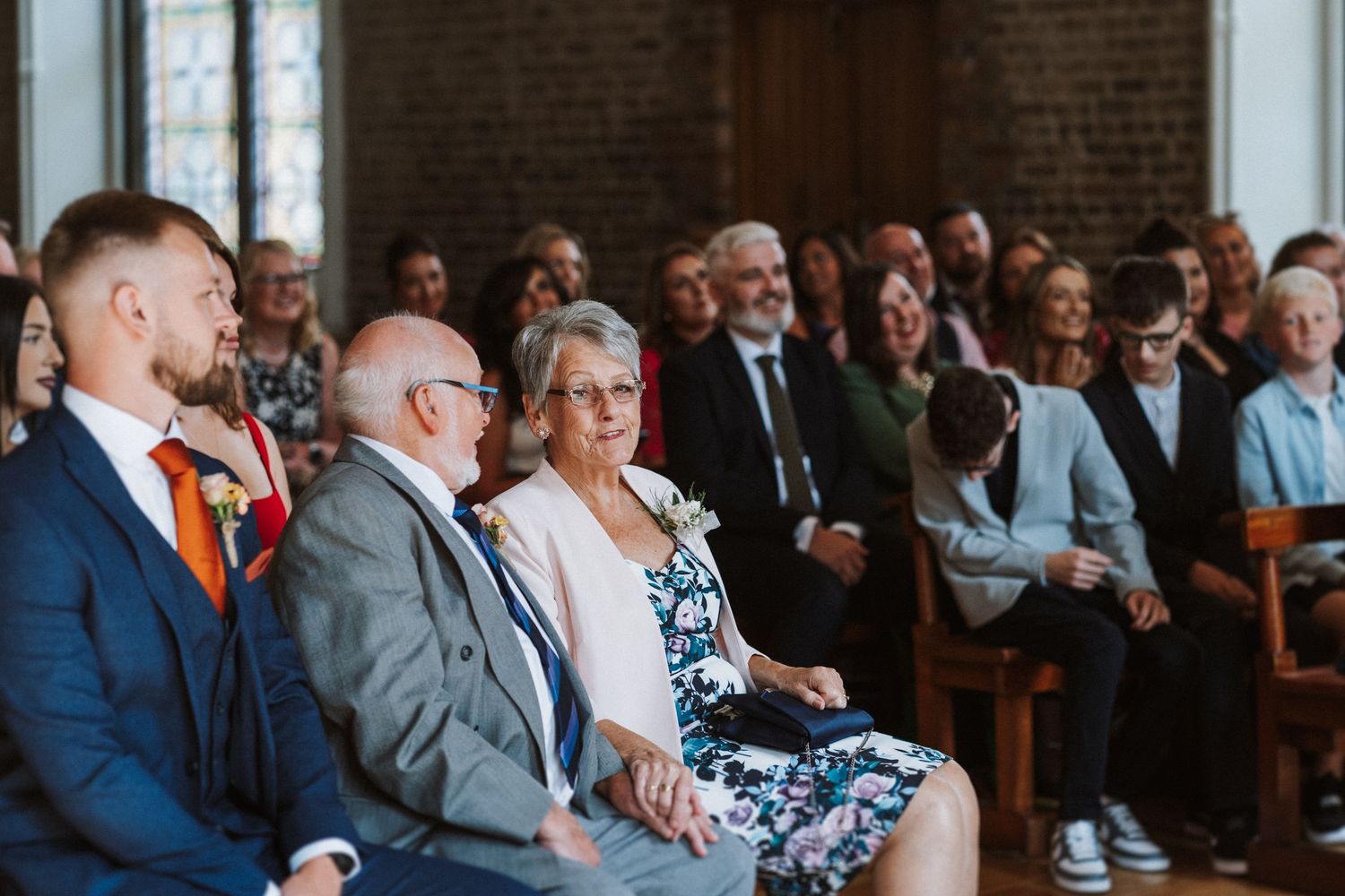 Wedding guests seated in church pews watch attentively during a ceremony in a brick-walled sanctuary.