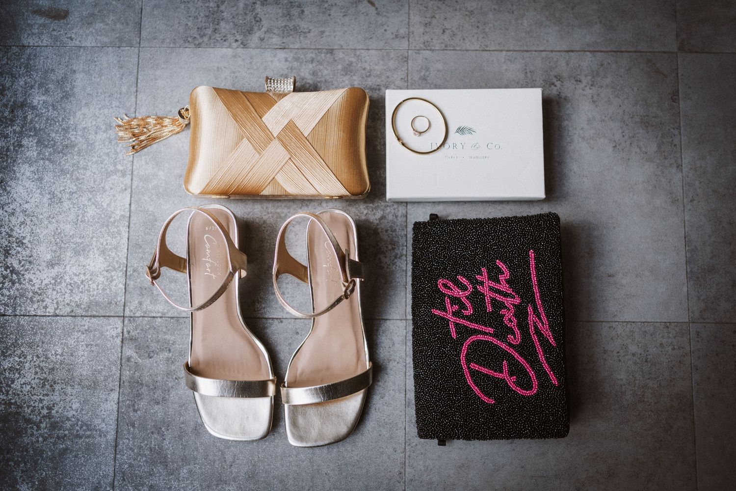 Wedding accessories including beige sandals, a metallic clutch, and monogrammed black towel laid on stone floor.