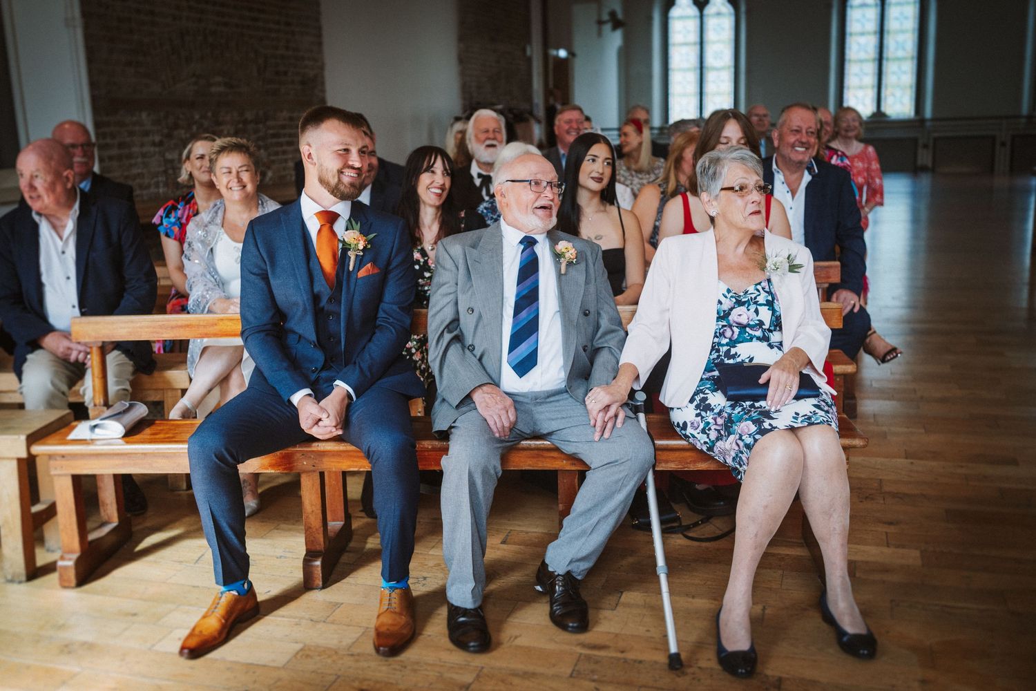 Wedding guests sit in church pews during a ceremony, with dress shoes and formal attire visible.