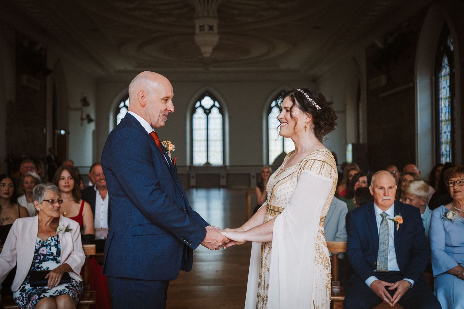 A wedding ceremony takes place in a historic chapel with gothic windows and gathered guests in attendance.