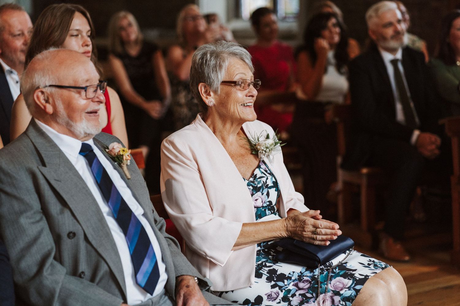 Audience members at a formal event share joyful reactions while seated in rows with brick walls behind them.