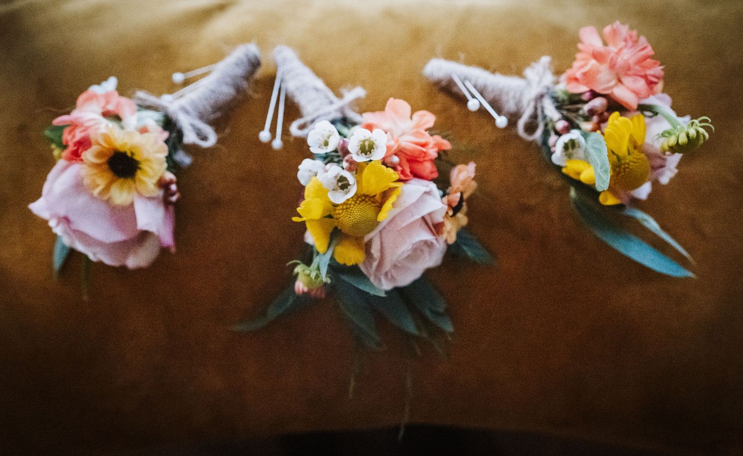 Three wedding boutonnieres with pink and yellow flowers and delicate greenery arranged on a brown surface.