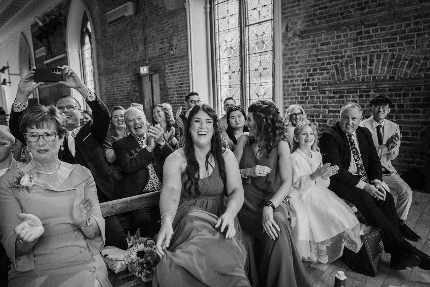 Wedding guests share a candid joyful moment while seated in church pews during a black and white ceremony photo.
