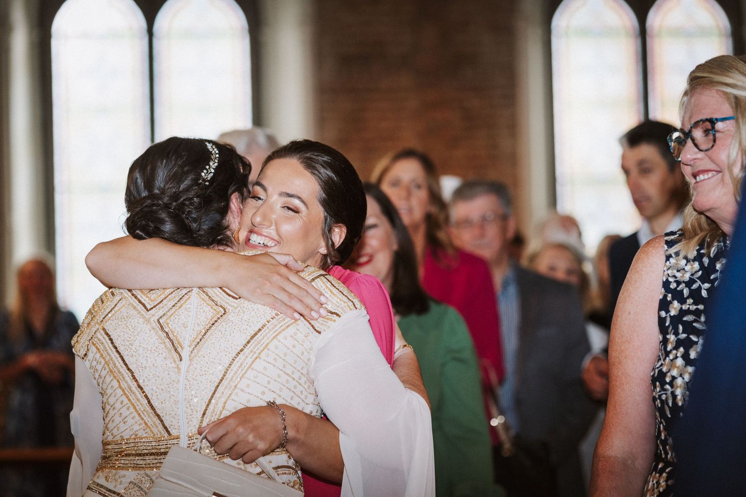 Two people share a warm embrace during an indoor celebration as guests look on in the background.