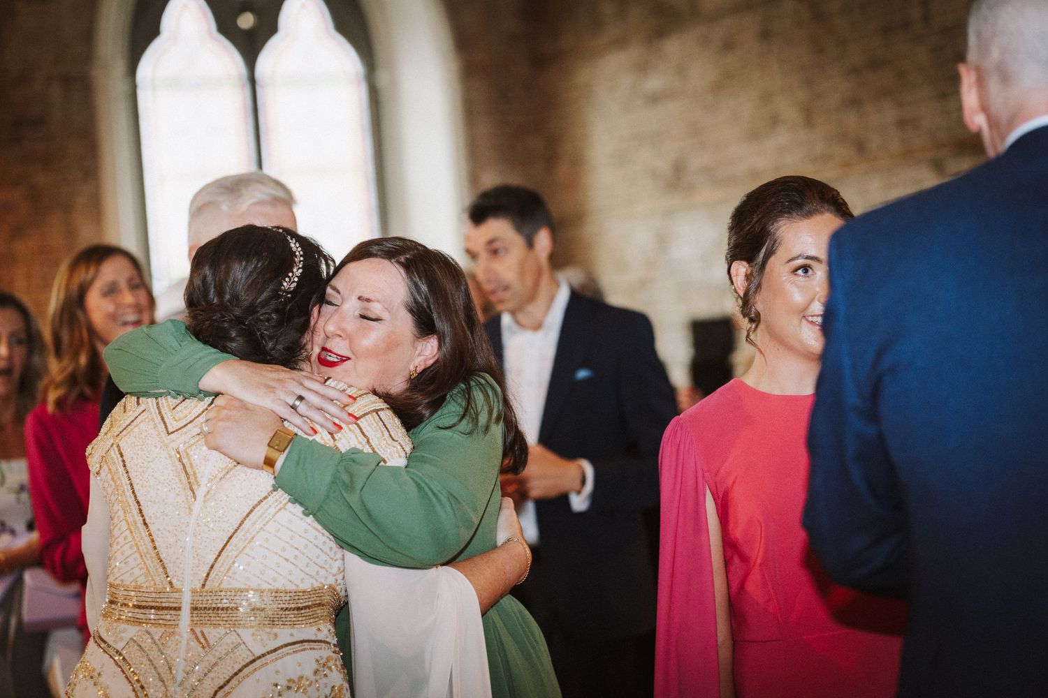Two people share an emotional hug at a formal event while other guests mingle in the background.