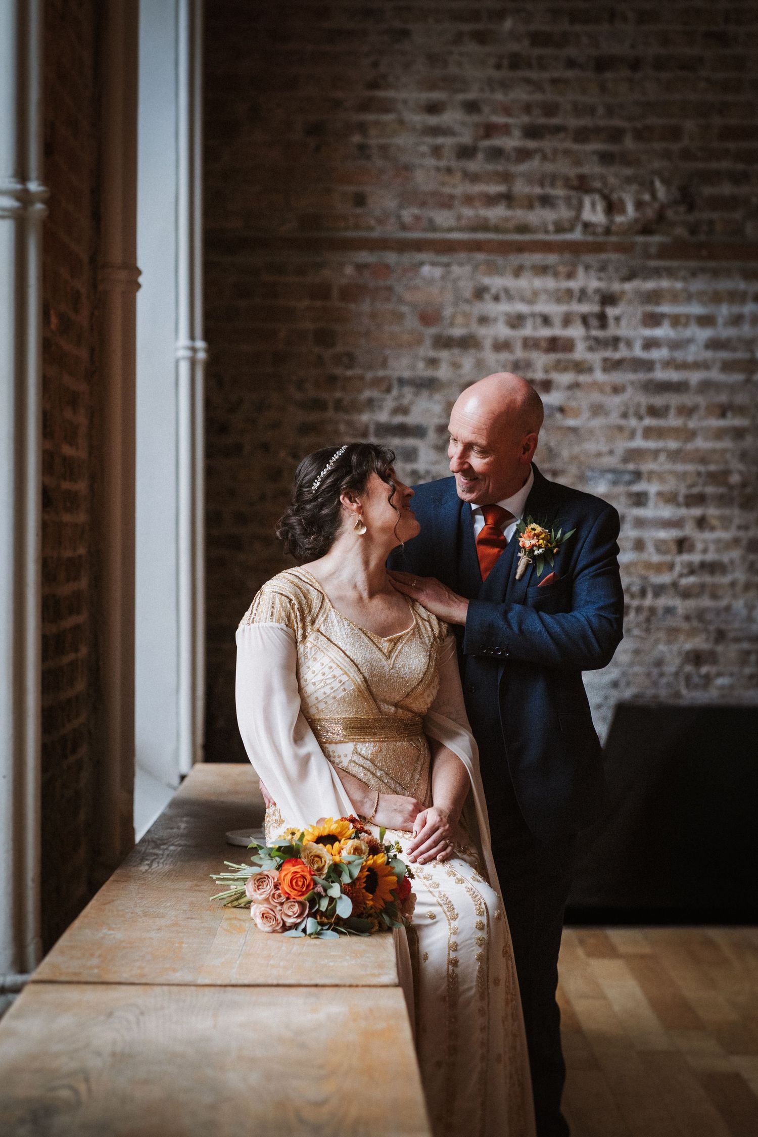 Couple in wedding attire embrace near brick wall, with orange floral bouquet on rustic wooden surface.