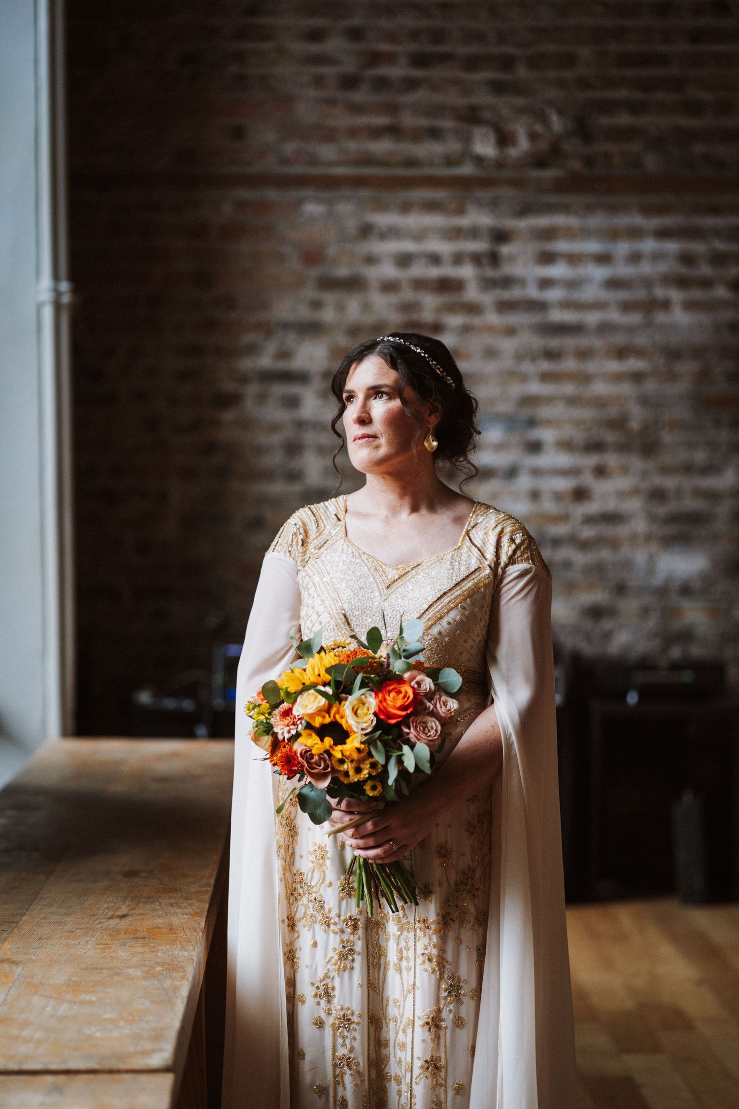 An elegant bride with curly hair wearing a vintage-inspired lace dress poses beside exposed brick walls in natural light.