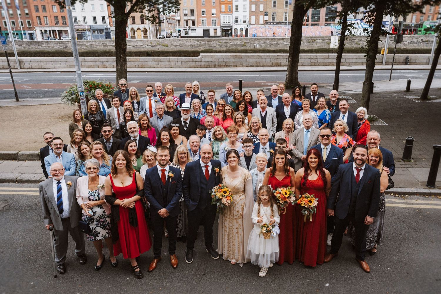 A wedding party group gathers for a photo in front of a stone building lined with autumn floral arrangements.