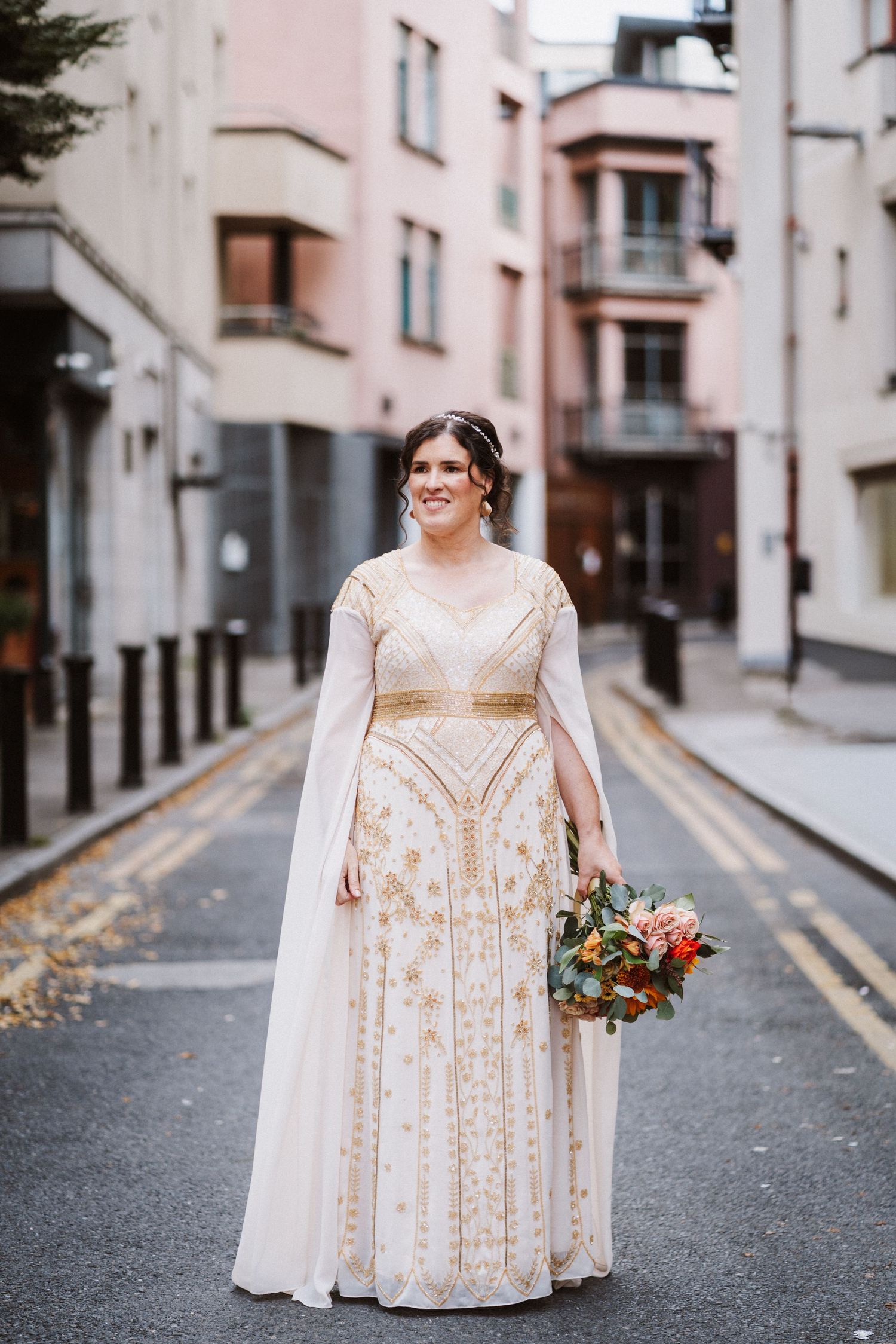 A bride in a champagne beaded gown poses in a charming urban alleyway holding a colorful autumn bouquet.