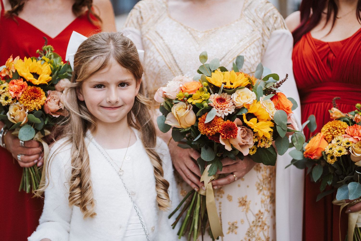Wedding party members hold vibrant autumn bouquets with peach, yellow and burgundy flowers against a white backdrop.