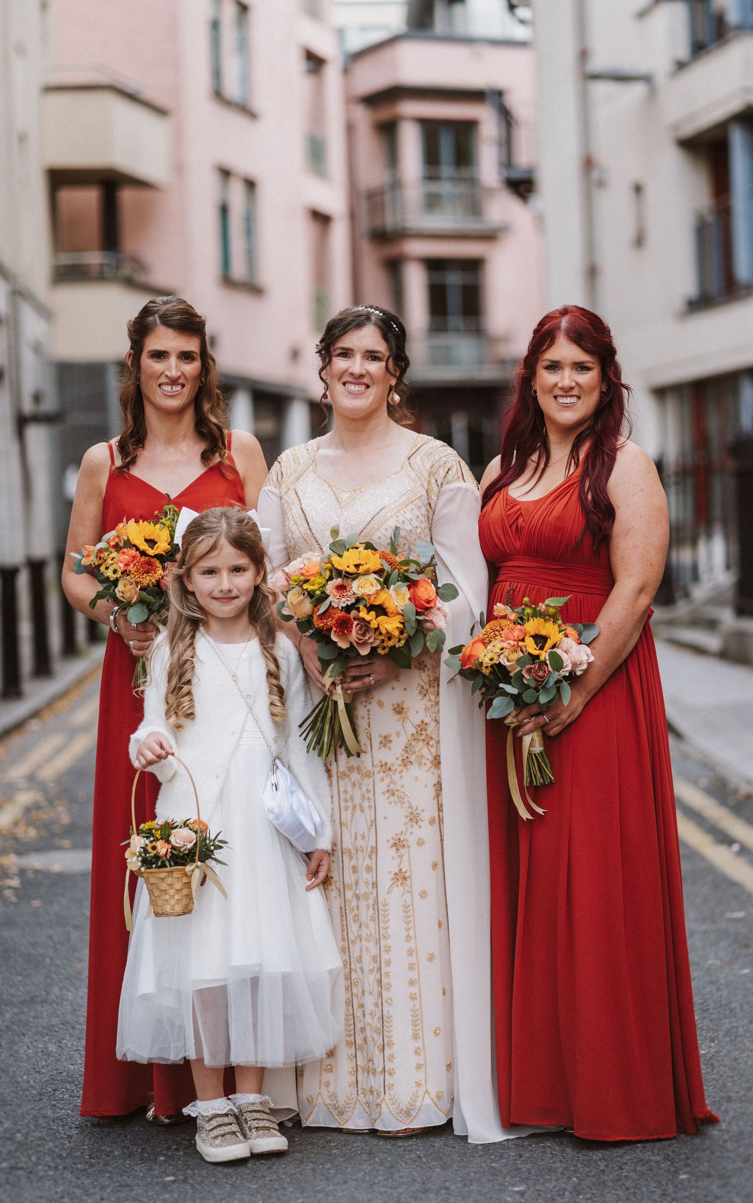 Wedding party members in red and cream dresses hold vibrant orange and yellow bouquets on a cobblestone street.