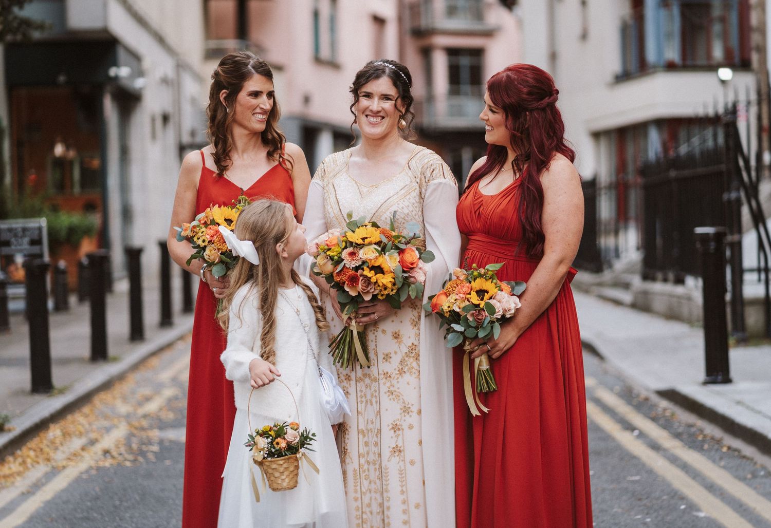 Wedding party wearing deep red bridesmaid dresses holding vibrant fall-colored floral bouquets on city street.