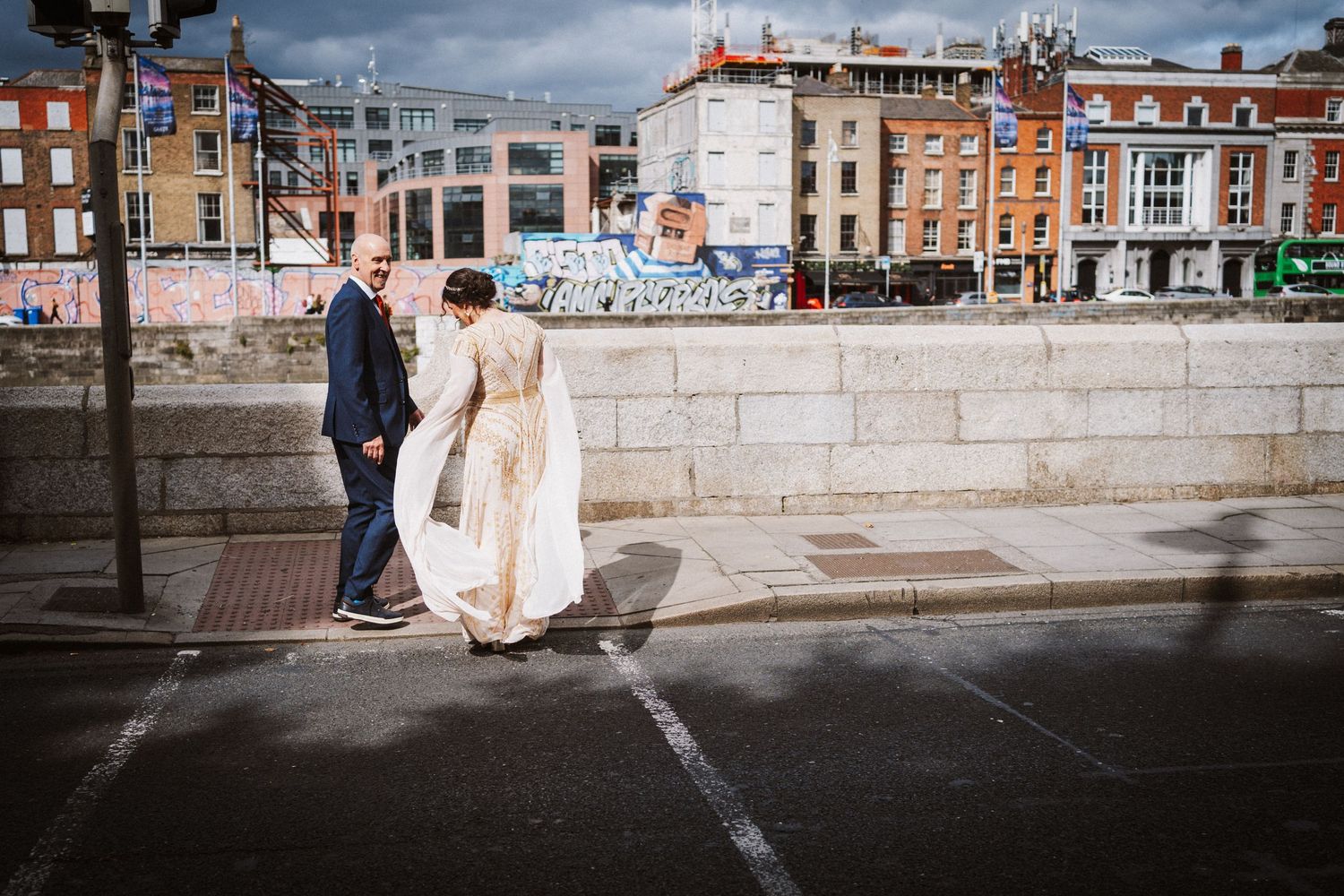 A couple dressed in formal wedding attire stands in front of stone walls on a sunny urban street.
