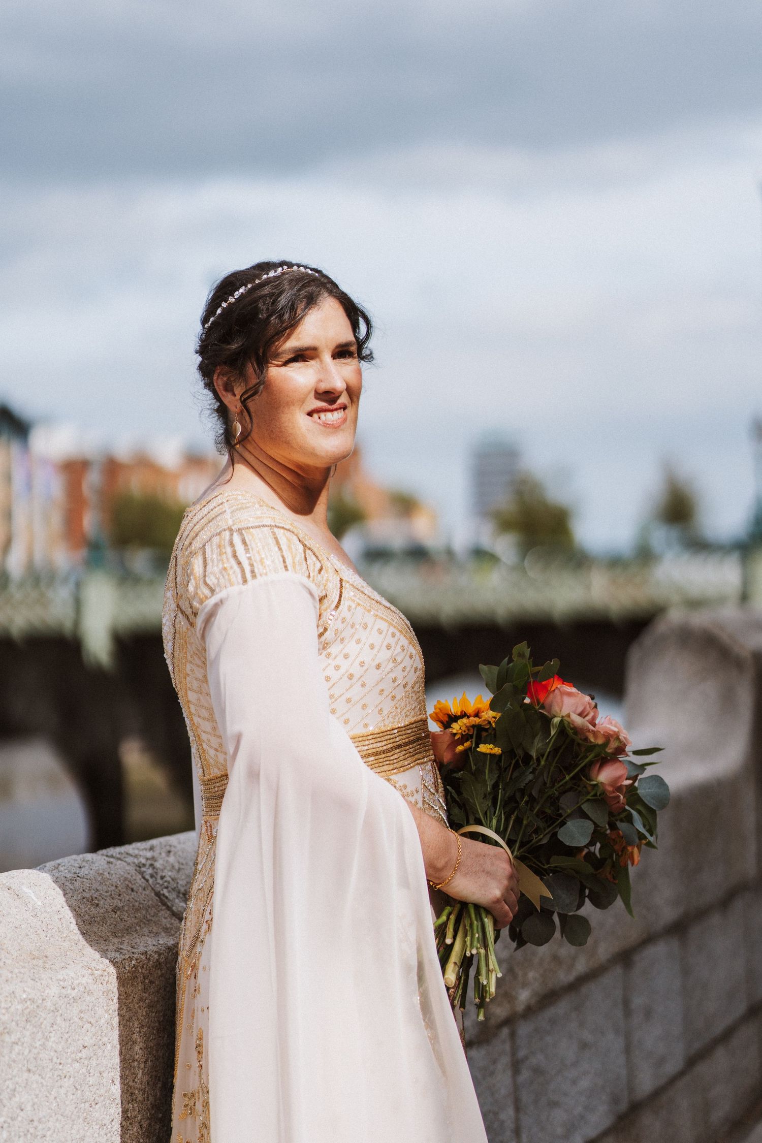 A bride in a flowing ivory dress holding an autumn bouquet poses along a stone wall on an overcast day.