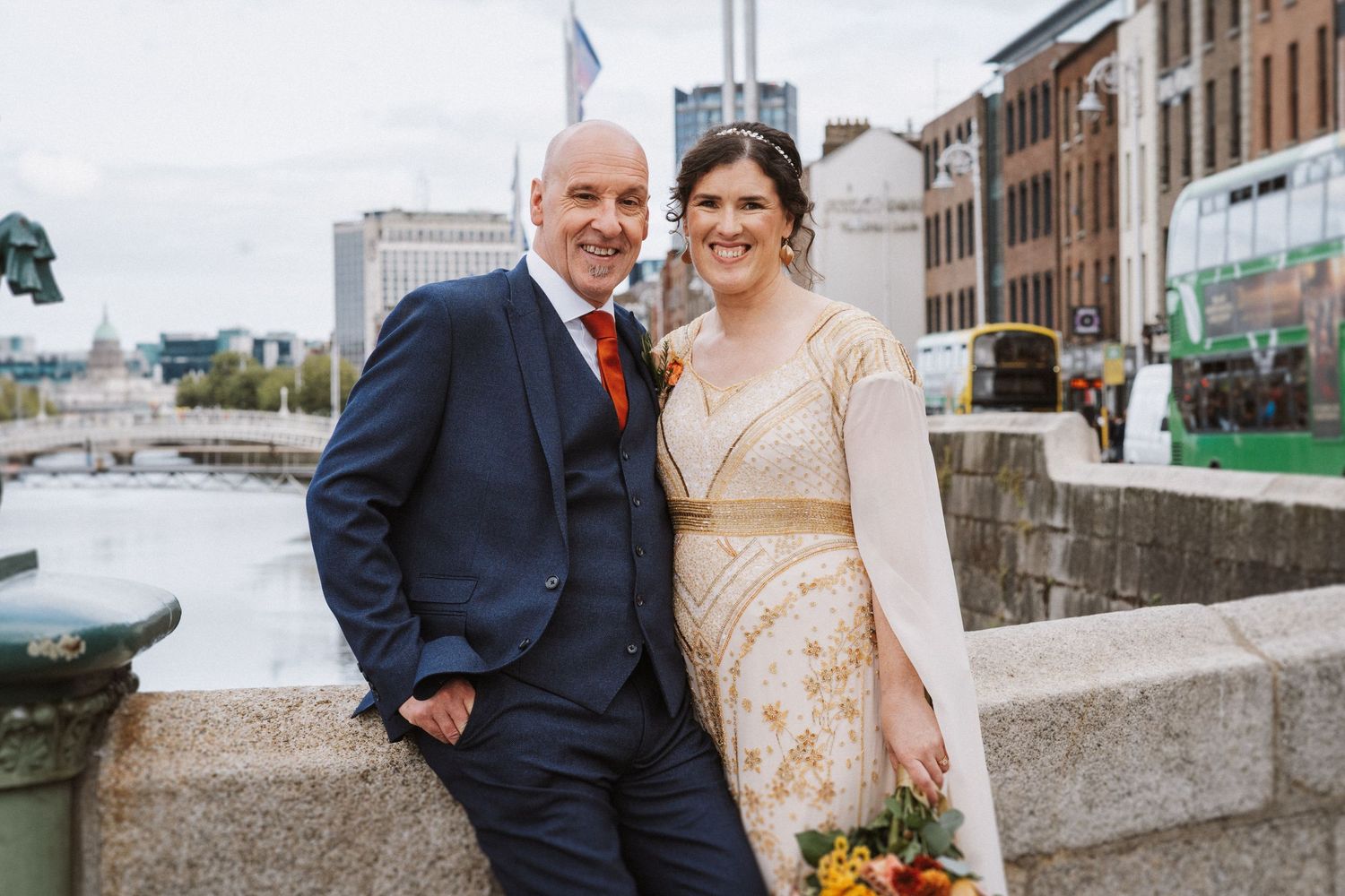 Newlyweds pose together on a stone bridge overlooking a river in an urban setting.