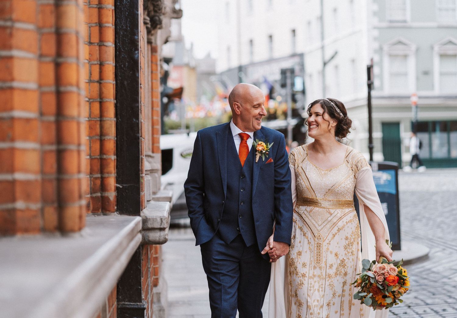 A couple shares a romantic moment on a cobblestone street beside an orange brick building.