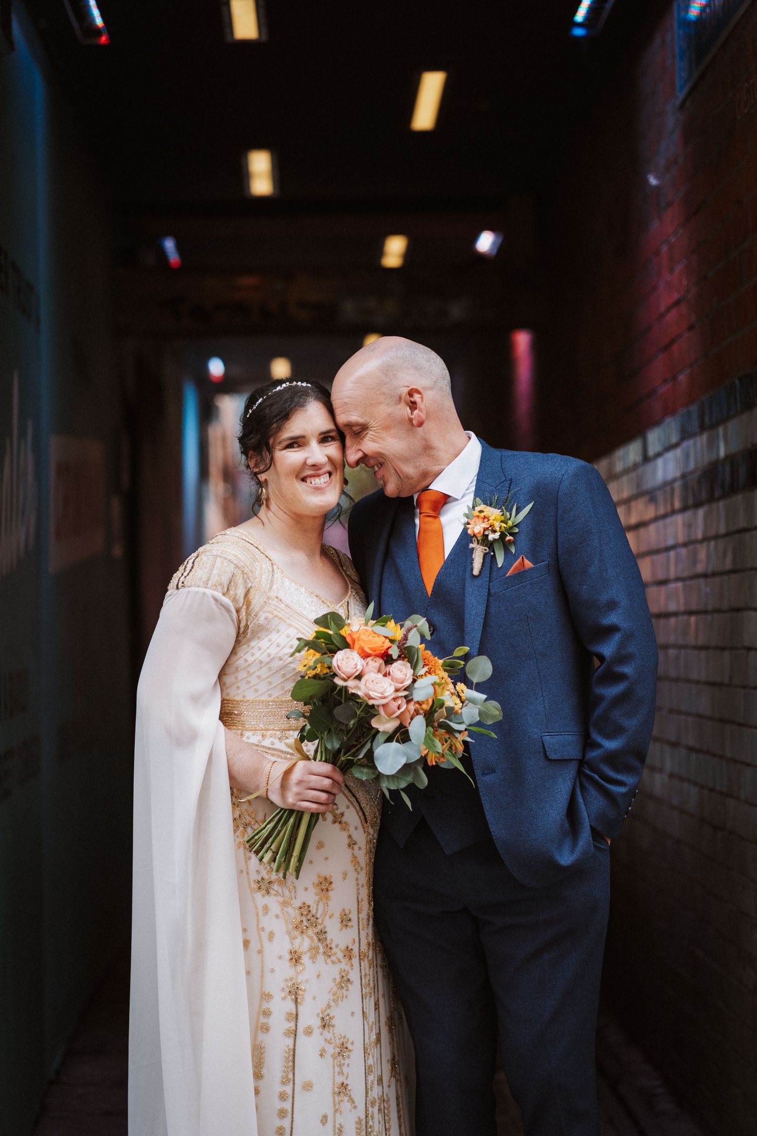 Bride and groom share intimate moments in dark brick hallway with orange floral bouquet and navy suit.