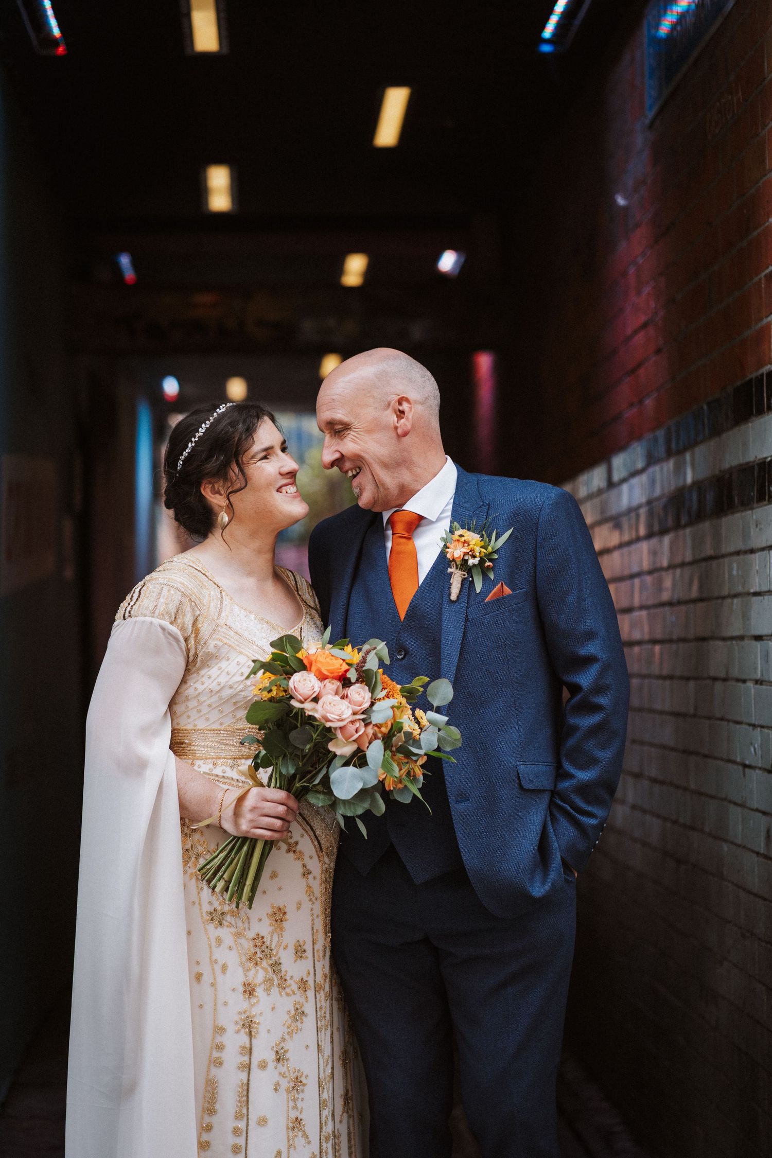 A tender wedding moment captured in dark hallway with couple in blue suit and gold floral dress holding autumn bouquet.