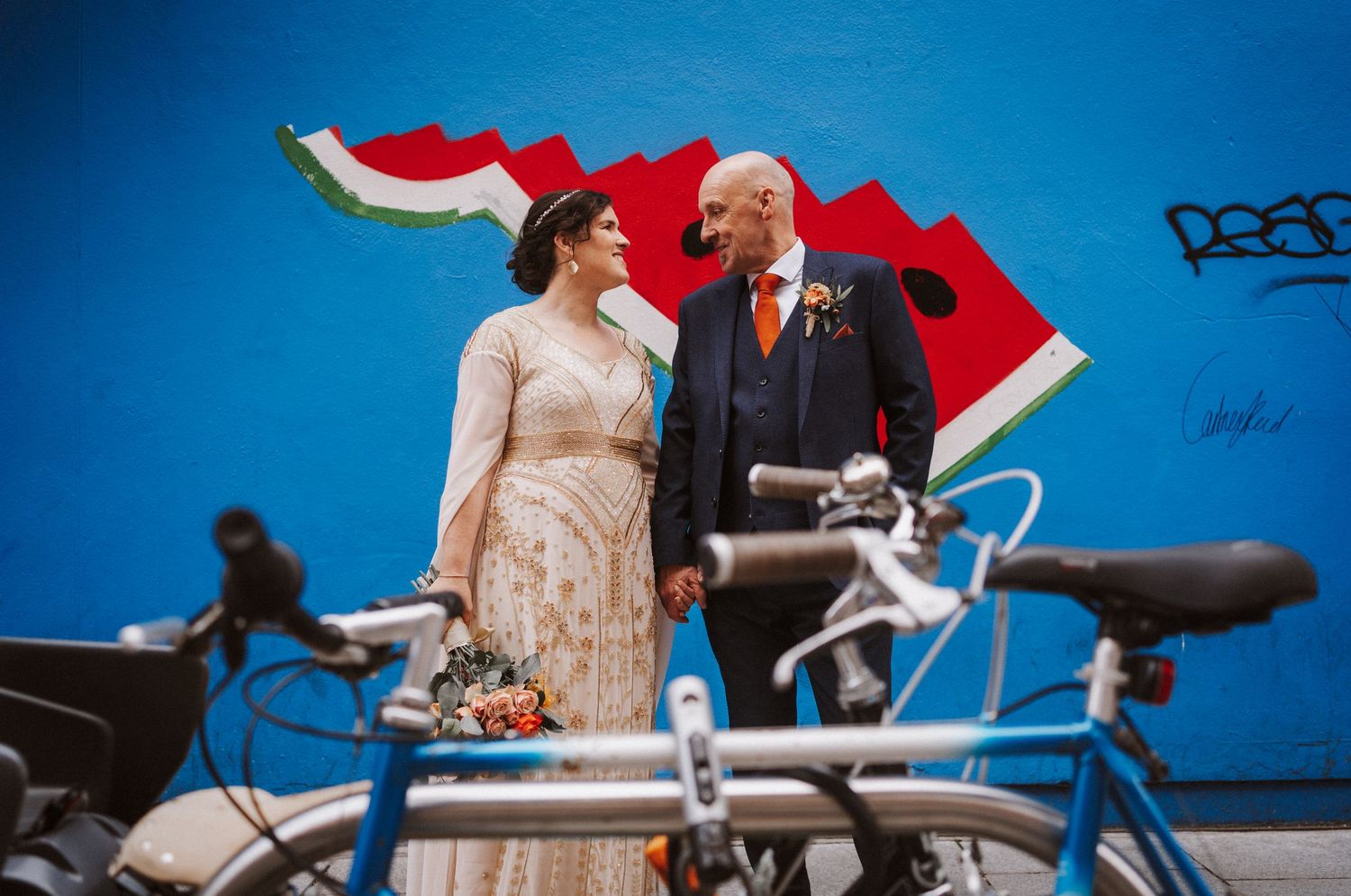 A couple poses with their red tandem bicycle against a bright blue wall, creating a vibrant urban backdrop.