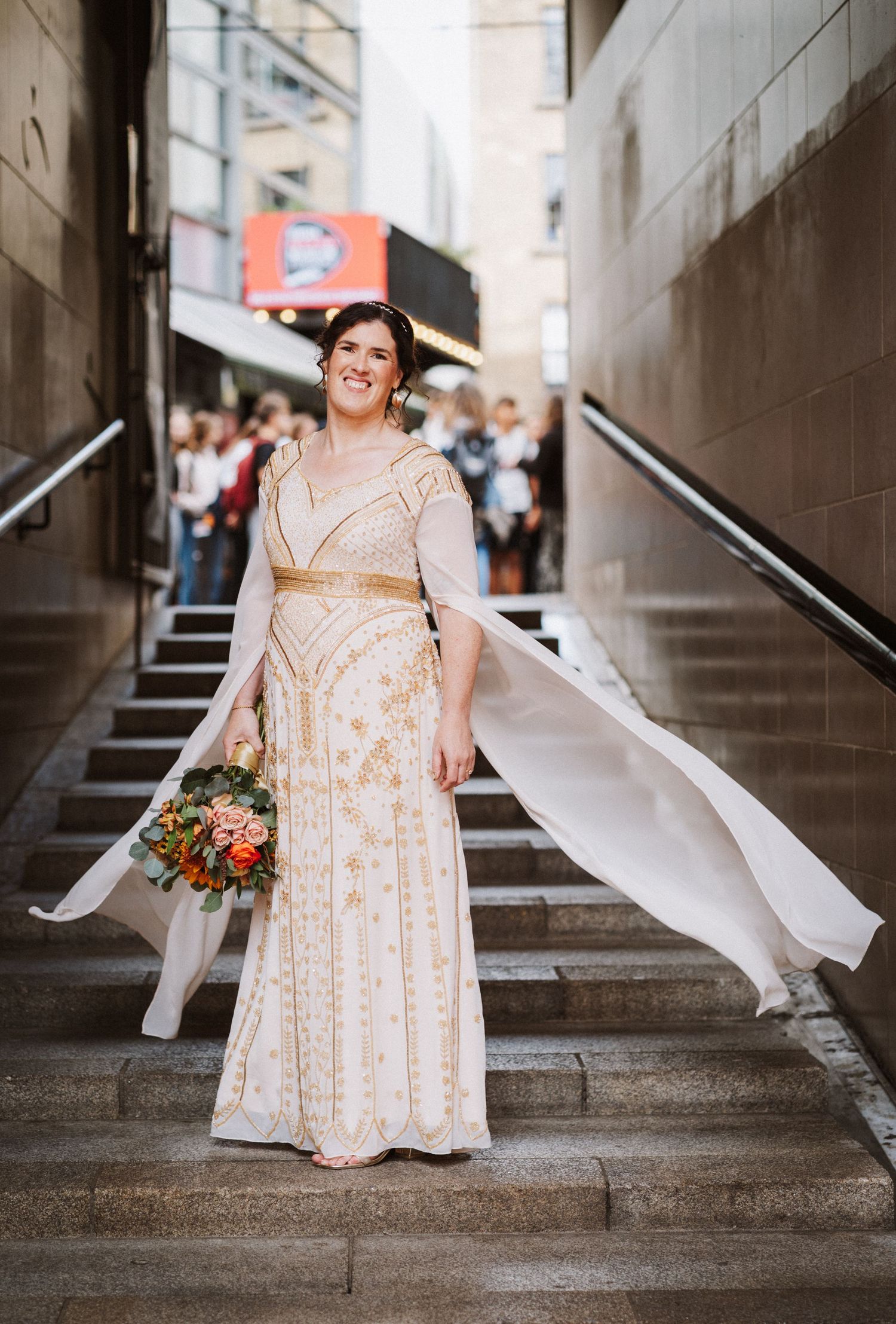 A bride stands on stone steps in radiant joy, dressed in a beige lace gown while flowing ribbons dance in the air.