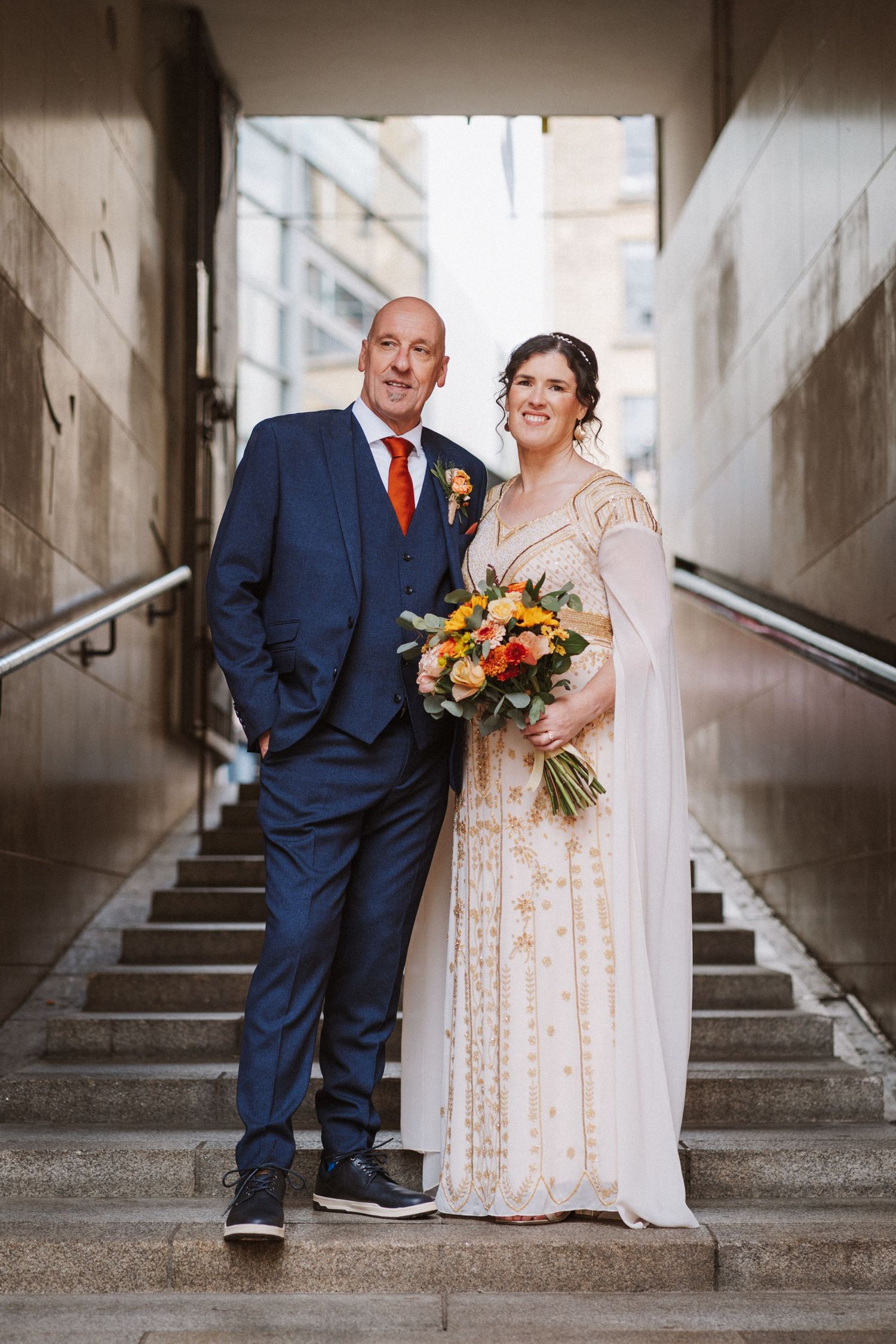 Bride and groom pose for wedding photos in a stark concrete stairwell wearing navy suit and cream lace dress with orange bouquet.