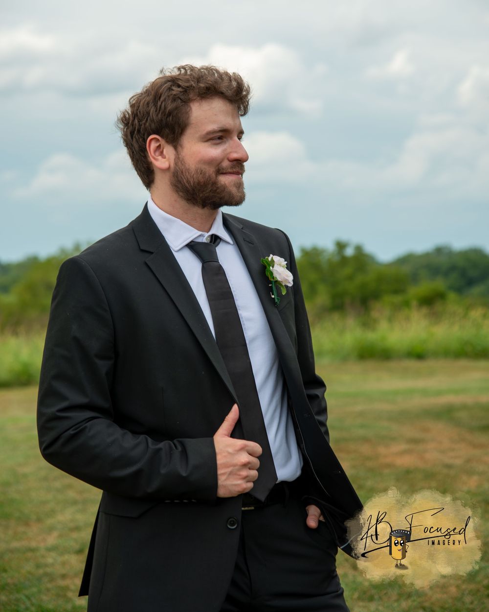 A person in a black suit and tie poses outdoors on a sunny day.