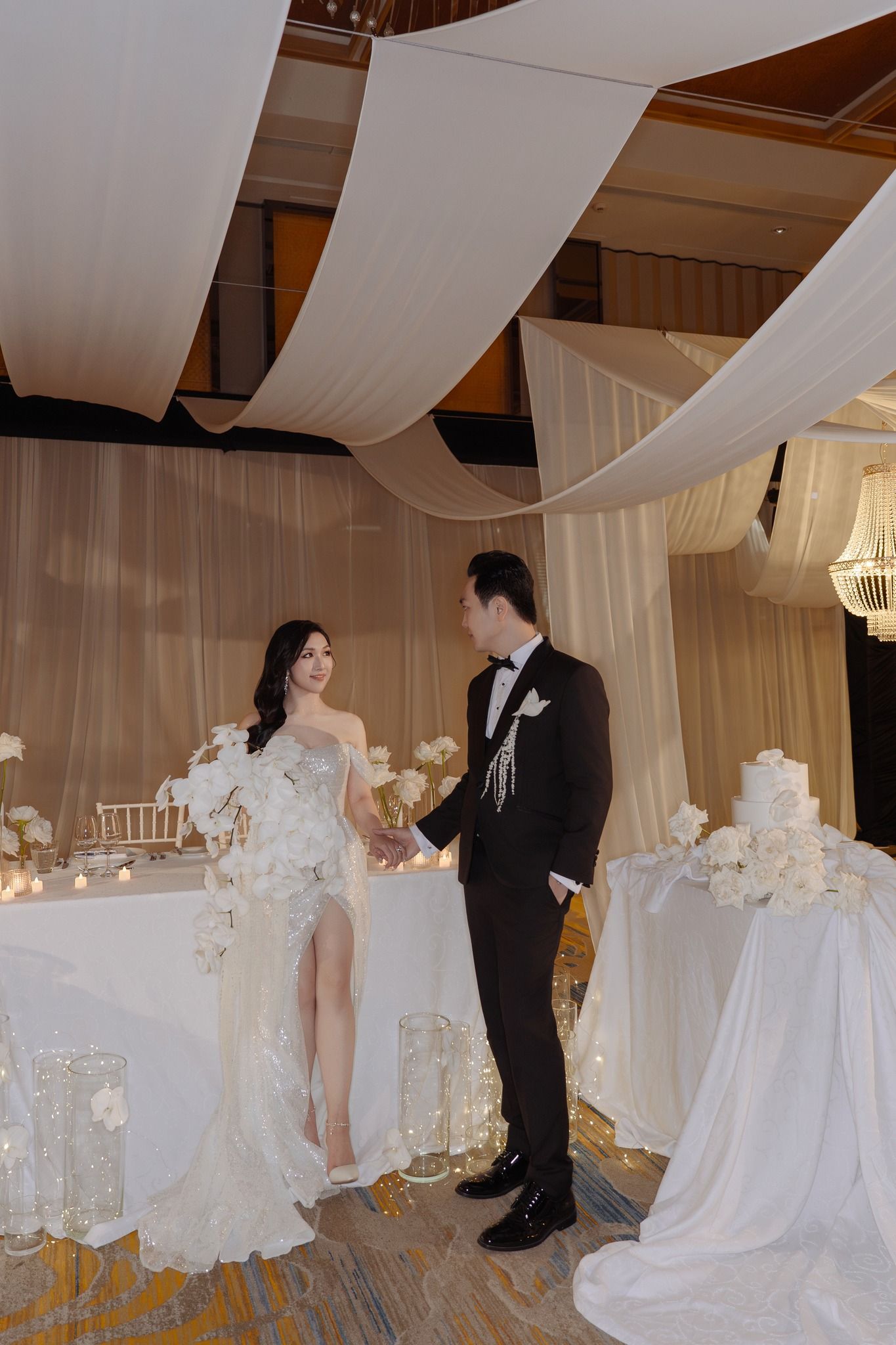 Bride in slit gown and groom in tuxedo holding hands at elegant white-draped wedding reception.