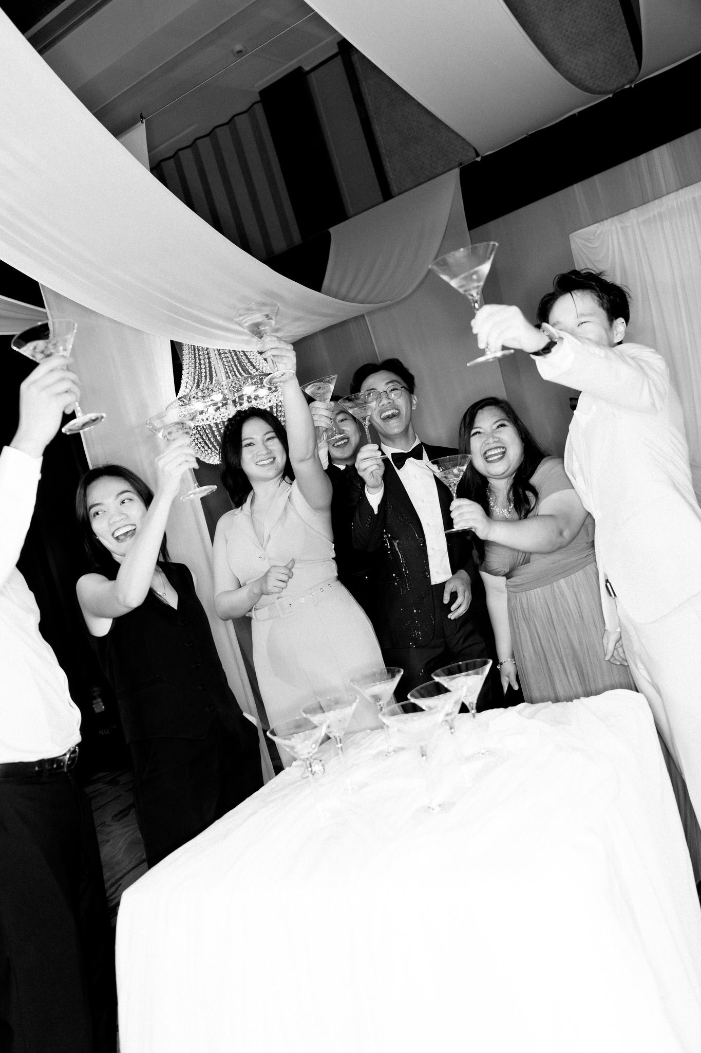 Black and white photo of wedding guests raising glasses in joyful champagne toast at reception.