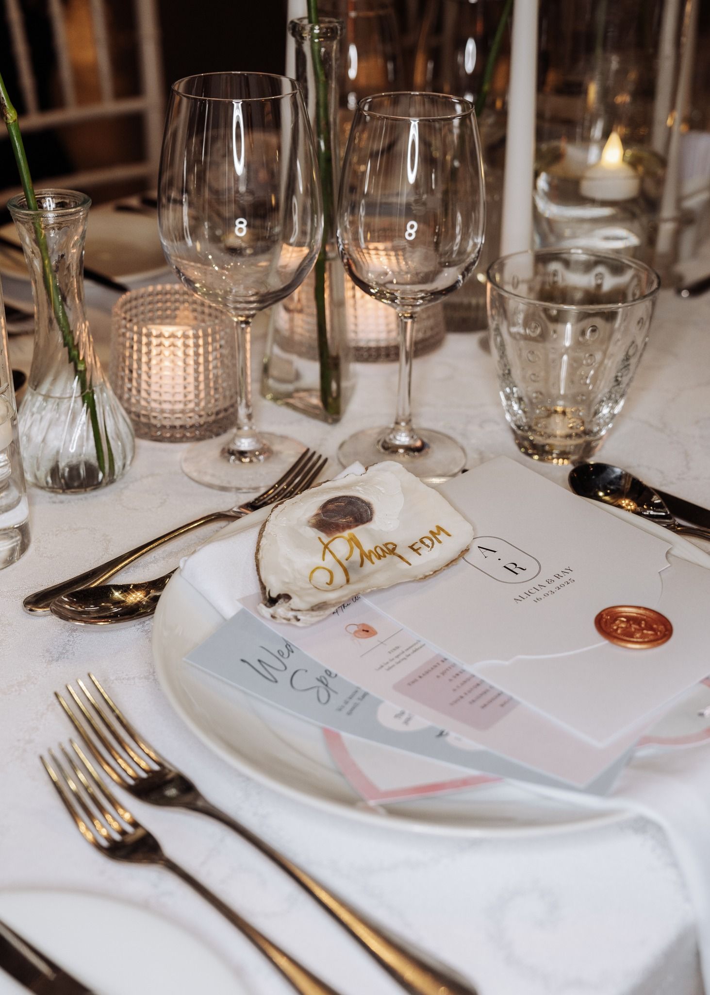 Elegant wedding place setting with gold cutlery, crystal glasses, menu card, and wax seal on white tablecloth.