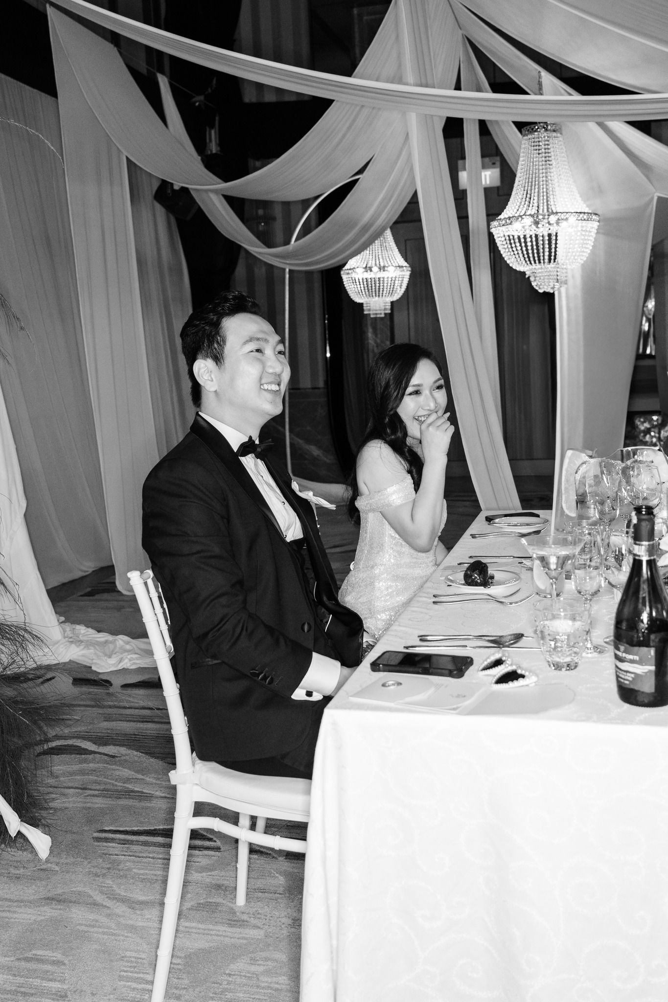 Black and white photo of smiling Asian couple at wedding reception dinner table with elegant draping.