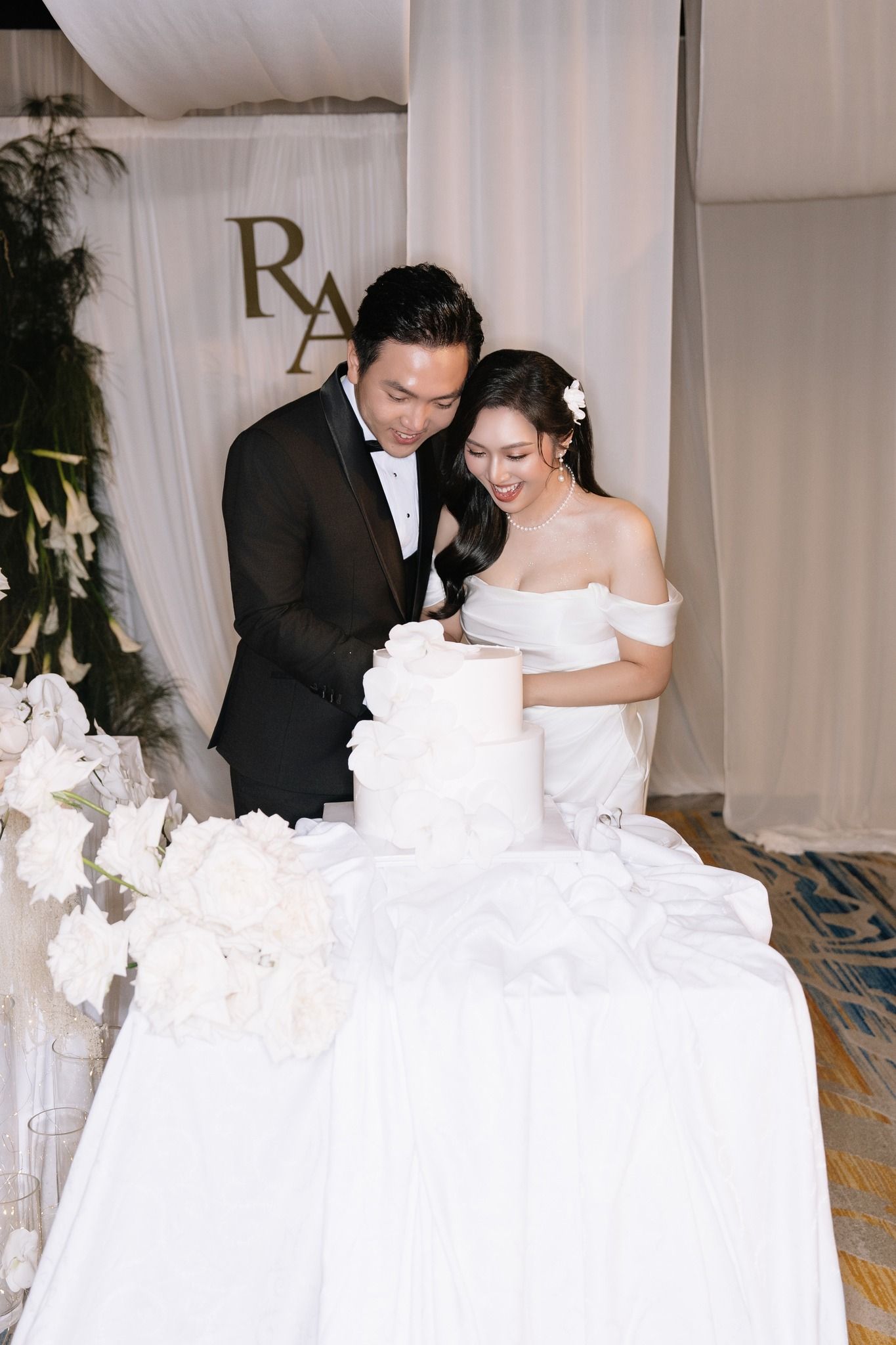 Asian bride and groom cutting white tiered wedding cake at elegant reception with monogram backdrop.