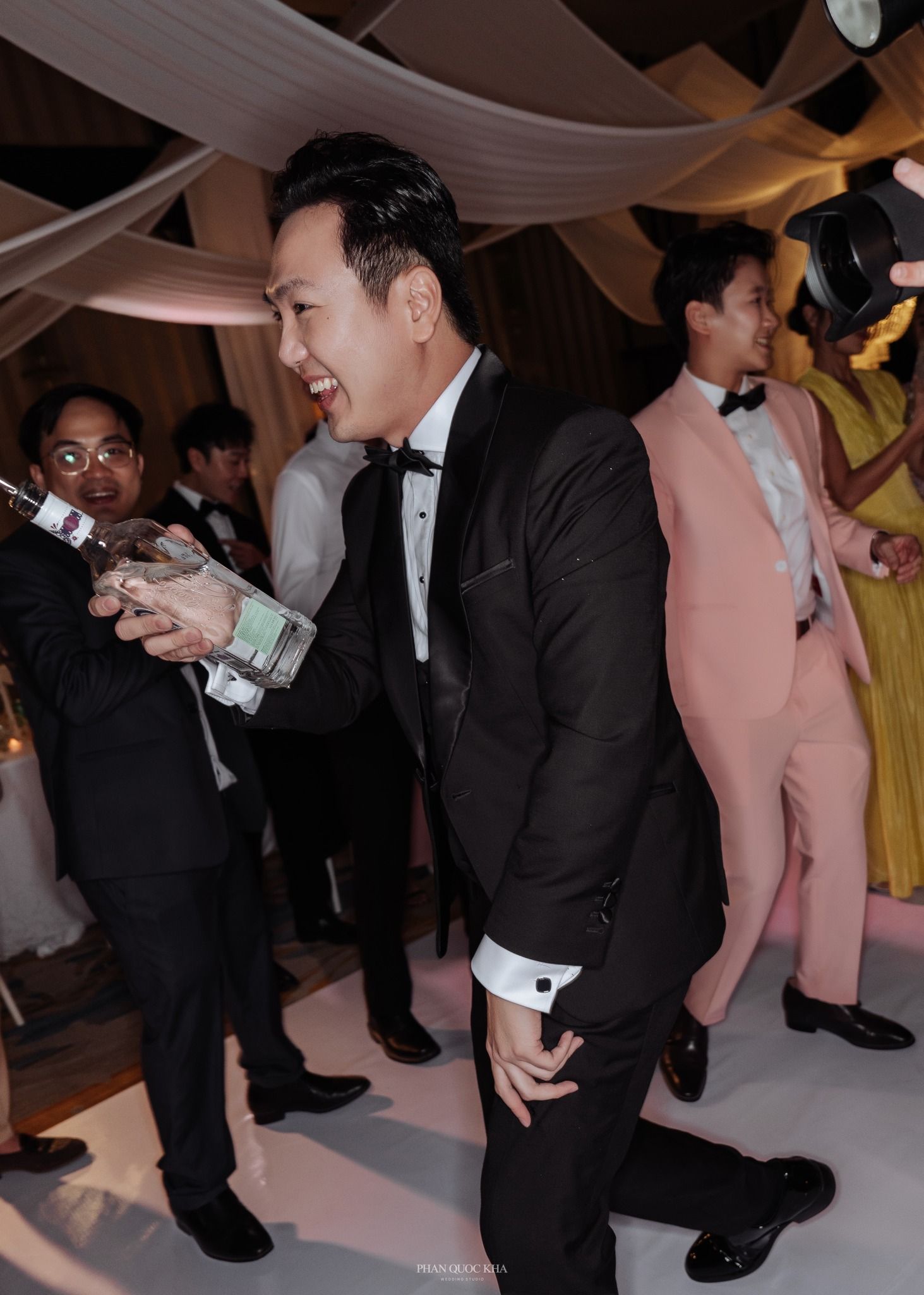 Laughing Asian groom in black tuxedo celebrating at wedding reception with guests in background.