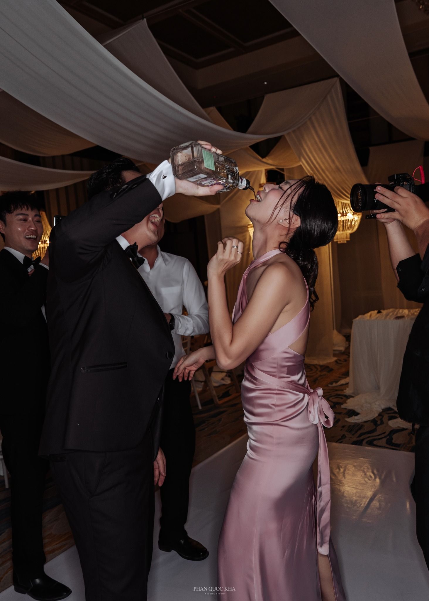 Wedding guests drinking from large bottle at lively reception party with draped ceiling decor.