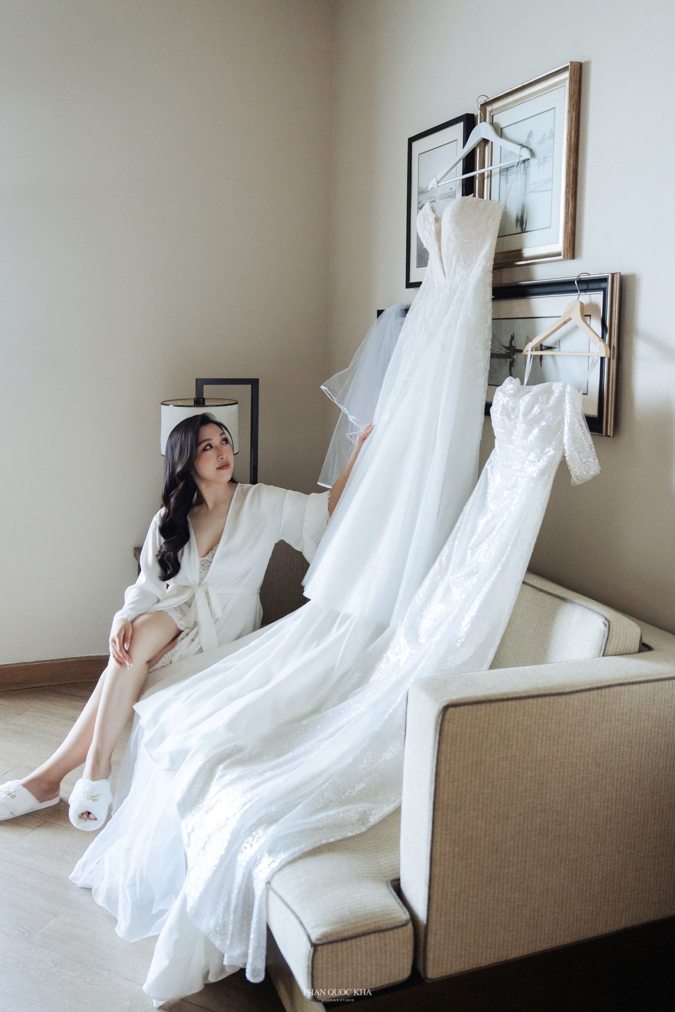 Bride in white robe sitting on sofa admiring hanging wedding dress and veil in hotel suite.